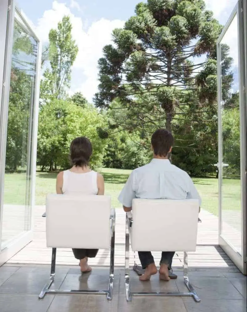 man and a woman sitting in white chairs looking outside to trees through glass doors